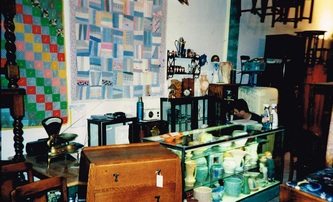 The first cash wrap at India Street Antiques after opening in October 1991. 

Quilts hang on the wall behind a glass case packed with art deco pottery.