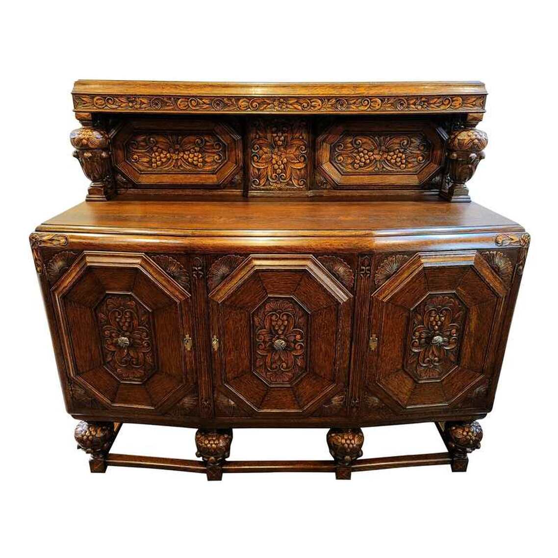 Post-1921 oak sideboard in an English Elizabethan court cupboard form. Court cupboard sideboards were popular in England in the 1920s and 1930s for displaying silver and pewter. The sideboard is very well constructed of solid oak wood. The top piece is removable for transport. The backboard features a flat top shelf with molded edge and a relief carved frieze of flowers, leaves, and scrolls. The back is enhanced with panels of relief carved flowers, leaves, scrolls, and grapes in octagonal framing. The stiles are enhanced with relief carved Art Nouveau flower buds with leaves. The cup-and-cover supports show relief carved acanthus leaves on the cup and grapes and leaves on the cover. The carcass has a center projecting geometric top with a softly molded edge. The top corners feature relief carved flowers and leaves. The front is fitted with three cabinet doors enhanced with octagonal framed panels of relief carved flowers, leaves, scrolls, and grapes. At the corners are relief carved anthemion. At the center of each door is a chased brass roundel pull featuring a ribbon