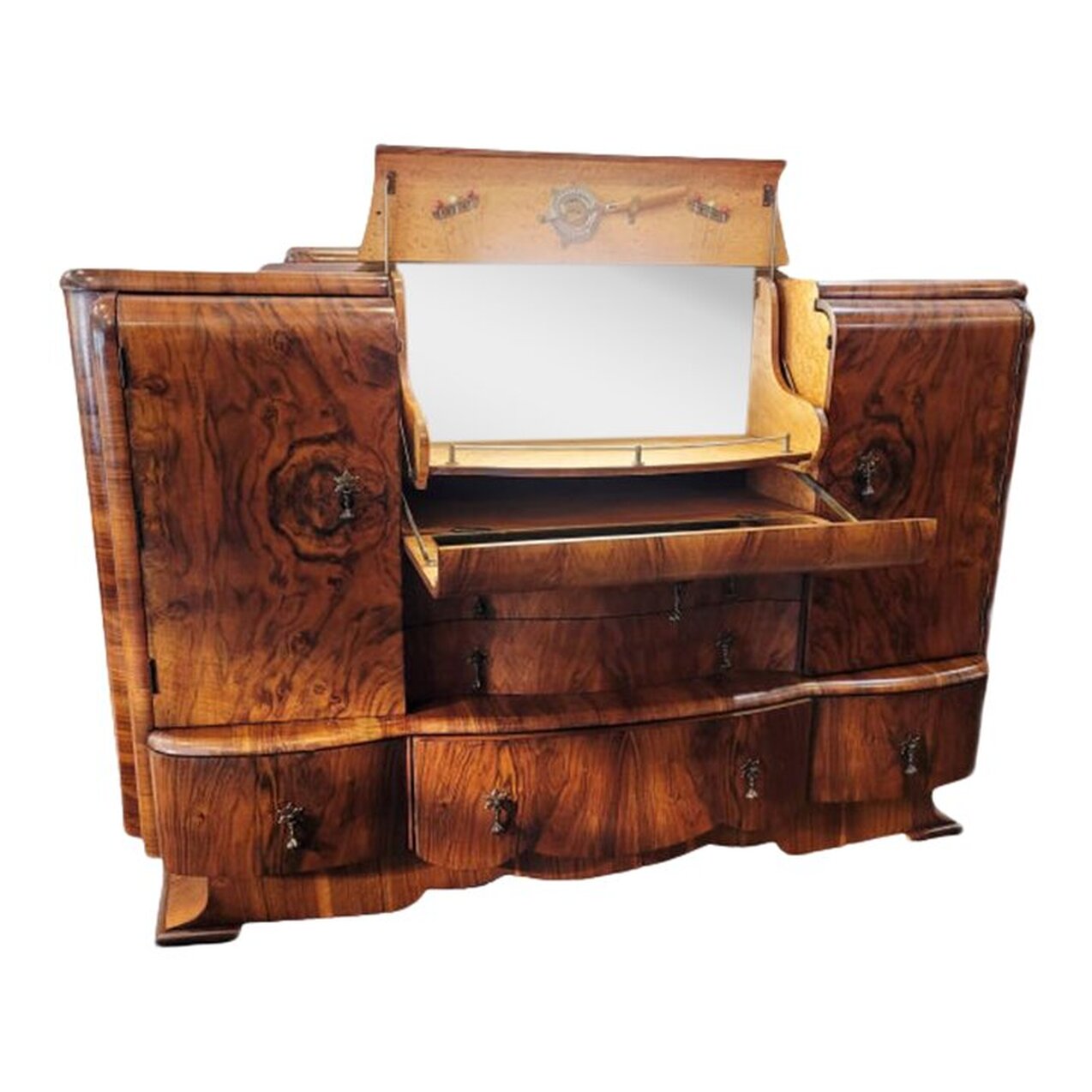 Vintage 1950s light-up cocktail sideboard was manufactured in England.  The carcass is finished with figured English walnut (Juglans Regia) veneers.  The shaped and leveled top is veneered with a waterfall figure. The back rail is carved at each end with a flower and leaf motif.  The middle top is fitted with a cocktail cabinet. The cabinet opens with a tug on the drop pull. The interior light will turn on when door is opened if left on. The light may be turned off with the press of the button on the front of the fixture. The interior of the drinks cabinet is lined with Birdseye figured Maple wood. Mirrors are installed on the back and on the inside of the lower cabinet door to be utilized as a prep station. Metal racks are installed inside the upper drinks cabinet door for a reamer or strainer and for cocktail picks. The shaped interior shelf has a chrome gallery along the front to secure bottles, decanters, and glassware.  The front of the sideboard is fitted with two single door cabinets - one on each side of the central drawers. The cabinet doors are veneered with oyster figured Juglans Regia and open via a tug on the drop pulls. The interiors are fitted with one standard shelf each with labels on the front edges. The left cabinet label reads 