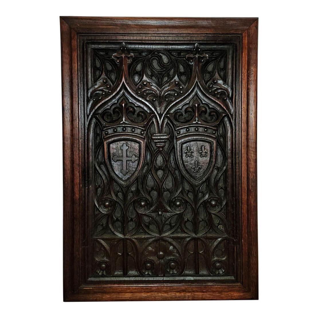 High Gothic tracery carved oak wood panel in oak frame.  Designs within the tracery include roundels, daggers, and cusps.  Two Gothic arches are carved over coats of arms. One coat of arms features a crown over a shield with a crosslet / cross bottony / cross trefly, or cross with trefoil tips. The second coat of arms features a crown over a shield with a trio of fleur-de-lis.
