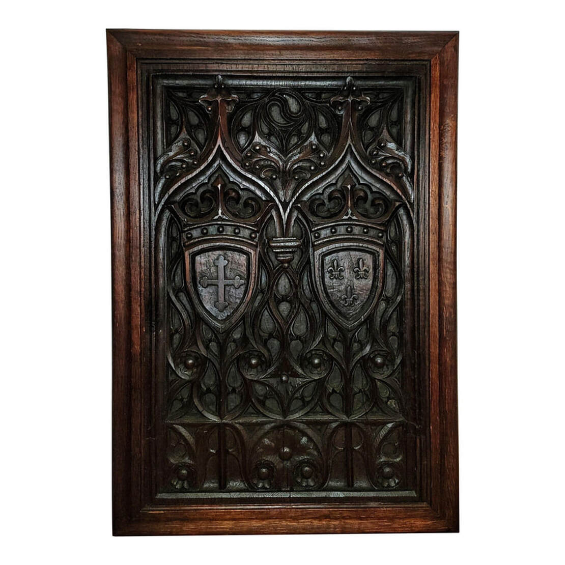 High Gothic tracery carved oak wood panel in oak frame. Designs within the tracery include roundels, daggers, and cusps. Two Gothic arches are carved over coats of arms. One coat of arms features a crown over a shield with a crosslet / cross bottony / cross trefly, or cross with trefoil tips. The second coat of arms features a crown over a shield with a trio of fleur-de-lis.
