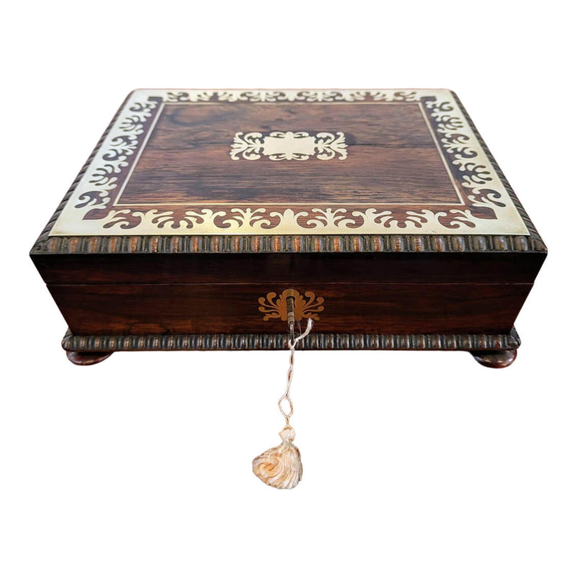 Antique Brazilian Rosewood canted rectangular box is decorated applied and inlaid brass work in the manner of Boulle. The box was created in the United Kingdom in the period 1795-1840 and most likely dates to the early 1820s. The lid features trim of applied foliated brass surrounding inlaid brass banding around a central inlaid foliated brass cartouche. Below the brass trim and at the base of the box is carved gadrooning. The box stands on turned rosewood knob feet. The box does lock and comes with the original key and silk tassel. The lock and the foliated escutcheon are brass. The interior lid is lined with striped moire silk fabric in pink and salmon tones. the interior base is fitted with cubbies for containing card, chips, and dice for gaming. The cubbies are lined with pink paper. The edges are gilded. The outer bottom of the box is lined with antique marbleized paper on which is printed page four of the fifteen-page 1752 Robert Roberts, Esq. book in which he sought to vindicate his actions as the creditor's agent in a lengthy and difficult process to settle the affairs of the post-late-1725 Irish banking business of Benjamin Burton, Samuel Burton, and Daniel Falkiner. This games casket witnessed many a fine card party! Box may also be used for jewelry and keepsakes. SIZE: 11