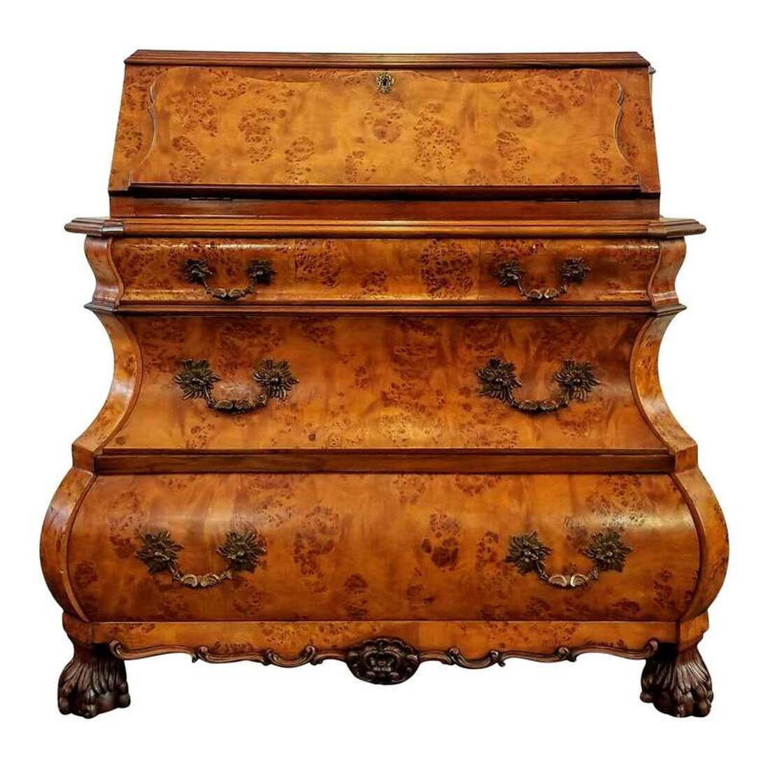 Dutch burr figure walnut wood bombe bureau in the Queen Anne / William and Mary style of the early 18th century. A cyma curve slant drop front secretary desk is below the shaped top and opens via chased brass hinges to reveal serpentine pigeon holes and two small storage drawers with turned brass knobs. The small interior drawer construction features a nod to the late-17th and early-18th century practice of placing the drawer bottom wood grain front to back. The desk does lock and comes with a key. The key escutcheon features a fan and scroll motif executed in repousse brass. Below that is a shaped top over cyma curve edged bombe three-drawer chest. The three shaped drawers open by tugging on the cast brass rocaille bail pulls featuring Flemish scrolls, fans, and fuchsia flower motifs. The bail pull brass scutcheons feature a stylized chrysanthemum motif. The carved apron at bottom features Flemish scrolls meeting in the center at a foliate cartouche. The four feet are carved in a mid-to-late Queen Anne period hairy lion paw and ball style. The back is an oak wood panel. The desk was made post-World War II, in the 1950s and is a recreation of early-18th century styles popular during the William and Mary, Queen Anne, and Early Georgian periods. The bureau is very well constructed. SIZE: 44
