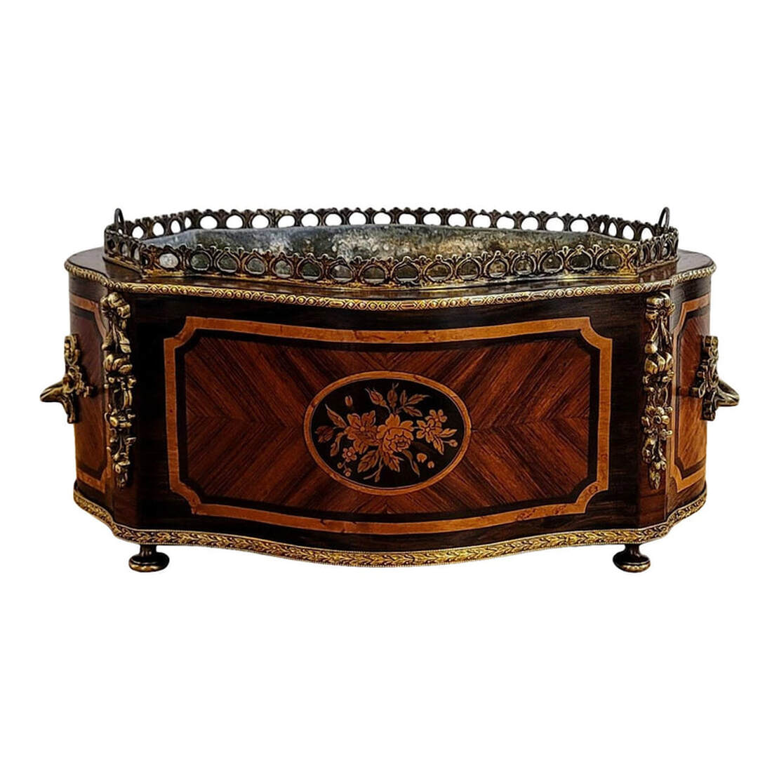 Antique jardiniere for tulips and orchids from the French period of Napoleon III, 1850-1870. The rosewood bombe shaped cachepot is decorated with veneer work in amboyna, yew, satinwood, kingwood, ebony and mahogany. The front and back feature a floral marquetry cartouche surrounded by amboyna banding, chevroned veneer, and all framed with ebony and amboyna banding. The sides are mounted with gilt bronze scrolled acanthus leaf handles over chevron veneer and framed with shaped banding in ebony and amboyna woods. On the top is a gilt bronze gallery pierced with a peacock tail eye design. The top and bottom edges are decorated with applied stamped decorative gilt bronze banding. The top banding is stamped with guilloche strapping. The bottom banding is stamped with a laurel leaf design. The four corners are decorated with gilt bronze ormolu floral pendants. The planter stands upon four turned gilt bronze knob feet. The jardiniere insert is bombe-shaped zinc with a ring pull on either side that folds down inside the insert.