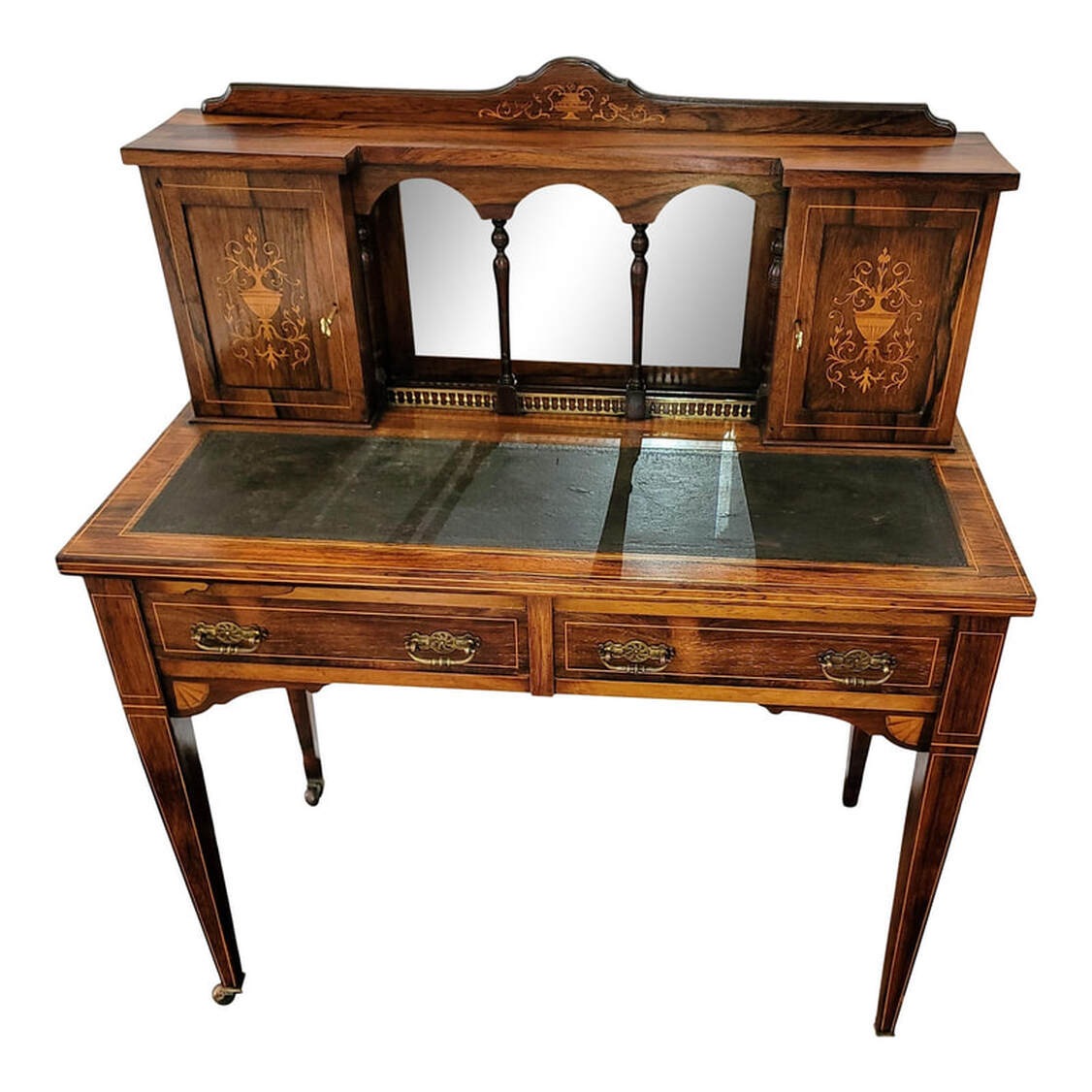 Late nineteenth century English rosewood writing table enriched with inlaid work by Maple & Company. The curved board across the top is inlaid at the center with a satinwood urn and scrolled acanthus leaves. The geometric shaped rosewood top rests upon two cupboards and an arcaded recess with a beveled mirror back, turned supports, and brass gallery. The cabinet doors are inlaid with satinwood urns and acanthus arabesques. The doors lock and come with keys. The left cabinet interior features two standard shelves and the right cabinet interior features three vertical pigeon holes for correspondence. The writing surface is inset with black leather embossed on the edges with a Greek key motif. Below the writing surface are two frieze drawers constructed with fine hand-cut dovetailing and hand-planed bottoms. The drawer pulls are a bail type on geometric Aesthetic Movement pierced and chased brass scutcheons. The square tapered legs end in brass casters. Inlaid satinwood banding accents the cabinet doors, the writing surface, the drawer fronts, the legs, and the desk edges. This model of writing table was sold by Maple & Company, England and was featured in their catalogue from 1885 to 1895.
