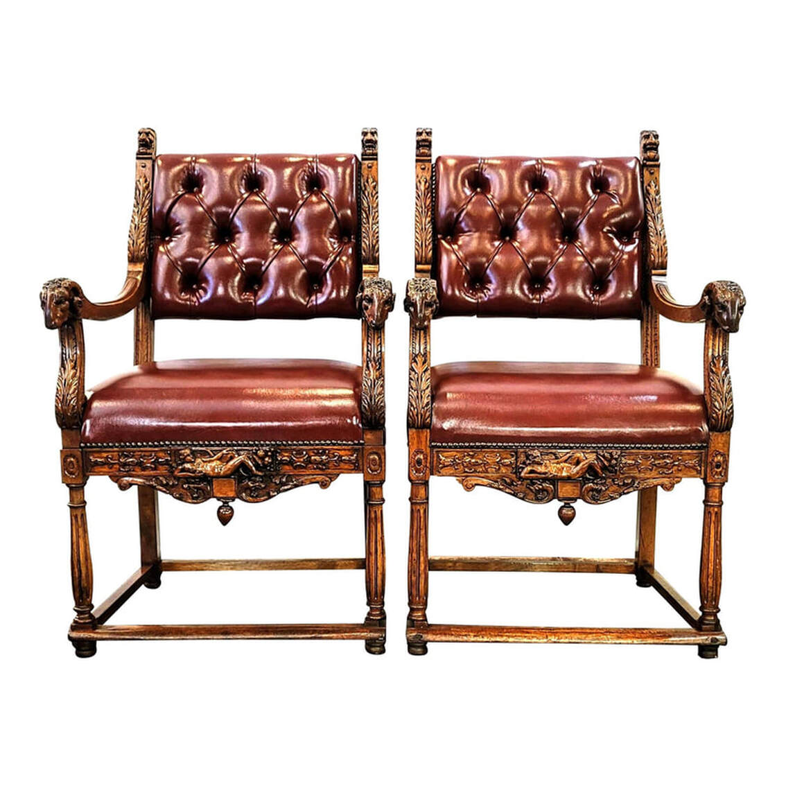 Pair late-19th-century through 1910 walnut French Renaissance Revival armchairs feature carvings of acanthus leaves, lion head finials, ram's heads at the ends of the arms, a nude reclining Demeter with cornucopia at the center of the front seat rails, and stylized scrolling dragons on the front aprons. The backs and seats are upholstered in leather-like burgundy fabric with button tufting on the backrests. Four low stretchers connect the legs above padded feet.