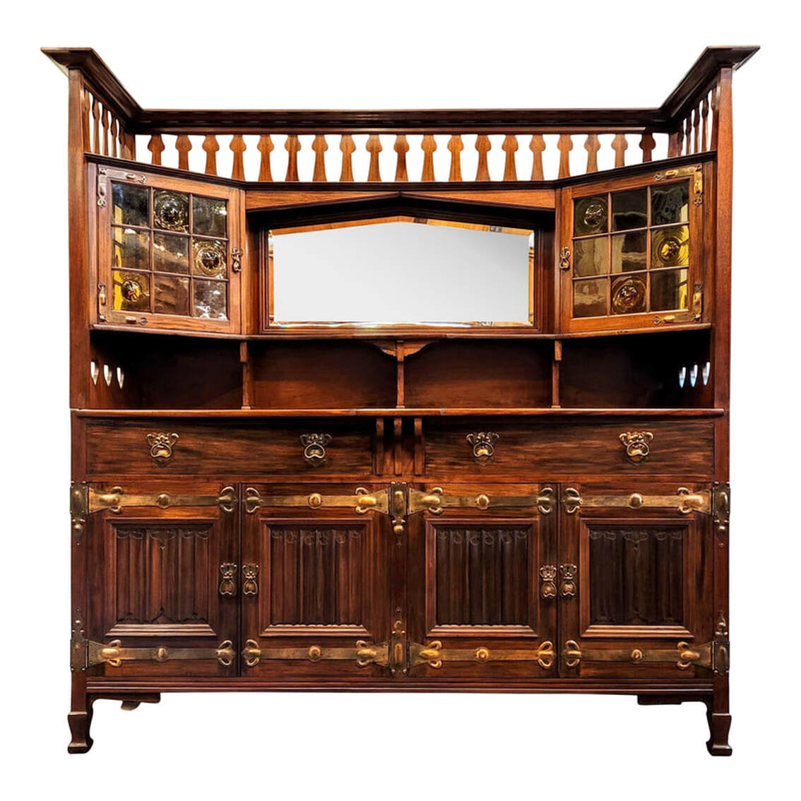Shapland & Petter main sideboard 2130 in fumed West Indies mahogany consists of a buffet carcase with cellarette and a mirrored over-mantle with glazed cupboards. The design of this sideboard dates from about 1894 through 1903. During this period, William Cowie was head of the design team which included James Henry Rudd and Henry Percy Shapland, grandson of the company founder. The over-mantle consists of a molded flat top shelf surround, double grooved for platter and plate display. Below this is a pierced gallery of peacock fretwork. The central shaped beveled mirror is inset into a pointed arch frame - a distinct Shapland & Petter treatment used virtually exclusively as a regular feature - surrounded by molding. Under the mirror is a shaped shelf. Below the shelf are three shaped support brackets. On each side is a canted cupboard with glazed doors and side panels. The glass panes are of a light amber-yellow tone. The plain panes are seedy glass and the remaining panes are blown crown antique bottle glass, or bullseye glass. The doors are decorated and attached with right angle corner pierced and hand-beaten repousse antiqued copper hinges. The drop pulls and pierced excutcheons are antiqued copper. The doors do lock and keys are provided. The inset brass lock on the left door is stamped
