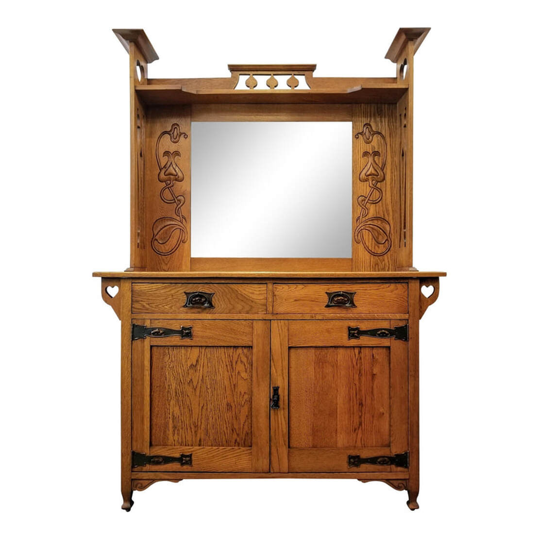 Antique main sideboard built with burl figured plain oak. The top is decorated with peacock tail fret rails above a shaped shelf. The side supports have William Cowie style heart cutouts at the top and Sheraton style urn fretwork throughout the rest of the supports. The central beveled mirror is framed on either side with oak panels into which are relief- carved Art Nouveau style bursting seed pods and flowers. The sideboard surface is above two frieze drawers fronted with single Art Nouveau / Arts & Crafts style copper bail pulls and back plates. To each side of the frieze are supports featuring the William Cowie syle cut-out hearts. Below the drawers are two cabinet doors with Arts & Crafts copper strap hinges and a single drop pull. The doors open to reveal a storage cabinet with a single shelf. The sideboard stands upon four spade feet. There are two curved apron brackets on the front.