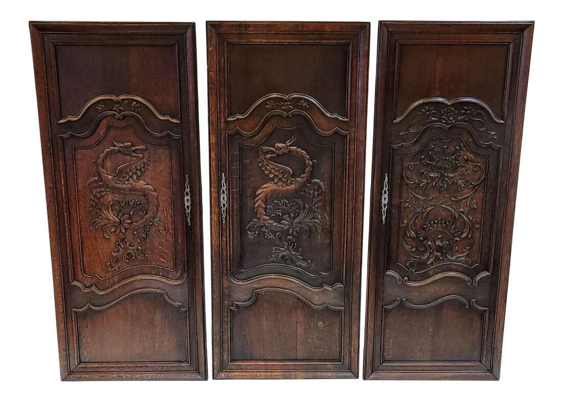 Three antique French oak armoire doors outfitted as hanging decorative wall panels.  Door panels date to the late-1800s and are in the French styles popular at the time of Japonisme, Gothic Revival, and Art Nouveau.  Two doors feature carvings of European style flying dragons in the manner of Gabriel Viardot, who created Japonisme furnishings in France after viewing the Chinese and Japanese furniture displayed at the 1867 Exposition Universelle in Paris.  The third door features carved foliated arabesques in rocaille style.  The carved panels are surrounded by scrolled frames.  The fronts retain the scrolled brass keyhole escutcheons.  The dragon panels measure 25.25