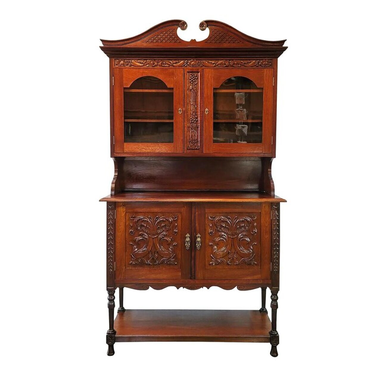 Solid walnut wood Georgian Welsh style cupboard on cabinet embellished with Neoclassical under the sea motifs of dolphins and shells is a fine example of upscale English Cottage furniture showing the Flemish influence on the vernacular furniture of South Wales. The molded crown is a sweeping broken swan's-neck pediment suggesting waves. The scrolls feature carved flower medallions. The front of the crown features relief carved fretwork. The frieze below the crown is bas-relief carved with dolphins and scallop half-shells with pearls. The upper vitrine is fitted with two removable wooden shelves and is fronted with two glazed doors. The wavy glass panes are square framed with a cyma curved top. The doors have solid brass locks applied to the interior and brass keyhole escutcheons in a scalloped Dutch style. The divider between the doors is bas-relief carved with acanthus stems and flowers sprouting from acanthus leaves and bundled with pleated ribbon. The upper side supports curve in just above the top of the lower cabinet. The backboard is plain with molded trim at bottom edge where it meets the top. The work top is flat with a molded edge. The lower cabinet is fitted with one fixed half-shelf and is fronted with two doors featuring inset panels bas-relief carved with dolphin pairs. The dolphins are classically presented with heads down and tails up. The fins and tails extend into foliated scrolls. The tails are tied together with pleated ribbon. The doors do lock via solid brass locks applied to the interiors. The keyhole escutcheons are a repoussé brass with a stylized dolphin motif reflecting the cabinet theming. The door pulls attached to the escutcheons are a molded brass open drop style. The cabinet apron features a curvaceously carved bottom edge with scrolls and a fan carved into the front. The lower cabinet side panels are plain with stylized s-curve aprons. The lower cabinet supports are bas-relief carved with half-flower motifs over long inverted acanthus stem pendants. A low shelf rests in between legs turned and ringed over blocks fronted with acanthus flower ornament. The feet are a turned wood elongated bun or ball style. The back is paneled in oak and walnut woods. 43.5