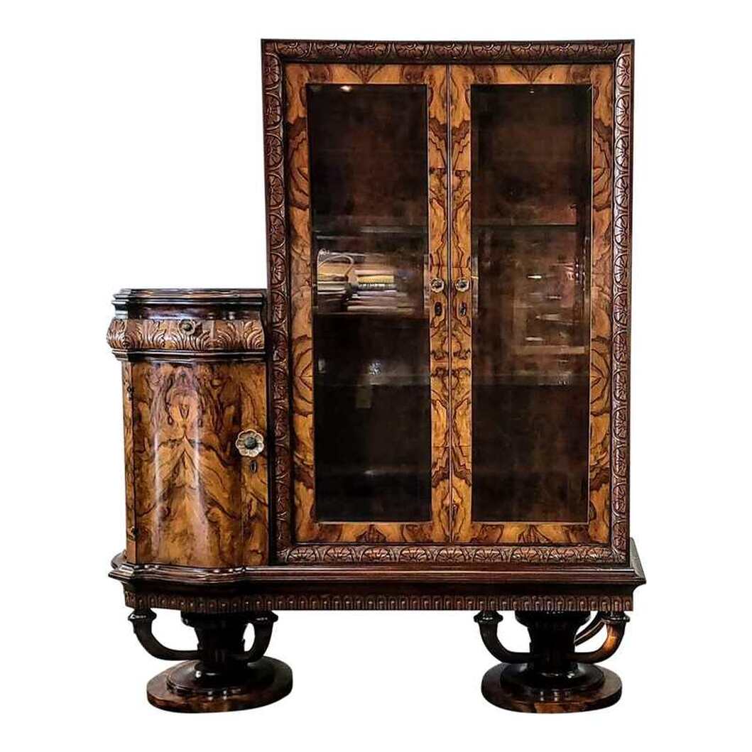 Vintage 1920s Italian Art Deco vitrine with attached storage drawer and cabinet veneered with matched burl figured olive wood and trimmed with walnut wood. The piece is well-built and finished with shellac. The large glazed display cabinet is placed to the right of a shaped cabinet with a storage drawer at top. The display cabinet has a flat rectangular wood top. The side panels are inset with beveled glass panes. The back of the interior is lined with burl figured olive wood veneer. Two glass shelves are provided for curio and china display; however, you may have wooden shelves made in order to hold books. The burled olive wood cabinet doors are inset with beveled glass panes. The carcase frame is executed in walnut wood and features carved floral lunettes with tree spacers of stepped Art Deco style. The doors do lock and a key is provided. The inset locks and latches are brass. The top latch is stamped